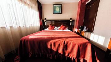 A hotel room with a large bed, a nightstand with a lamp, a window with curtains, and a painting on the wall.