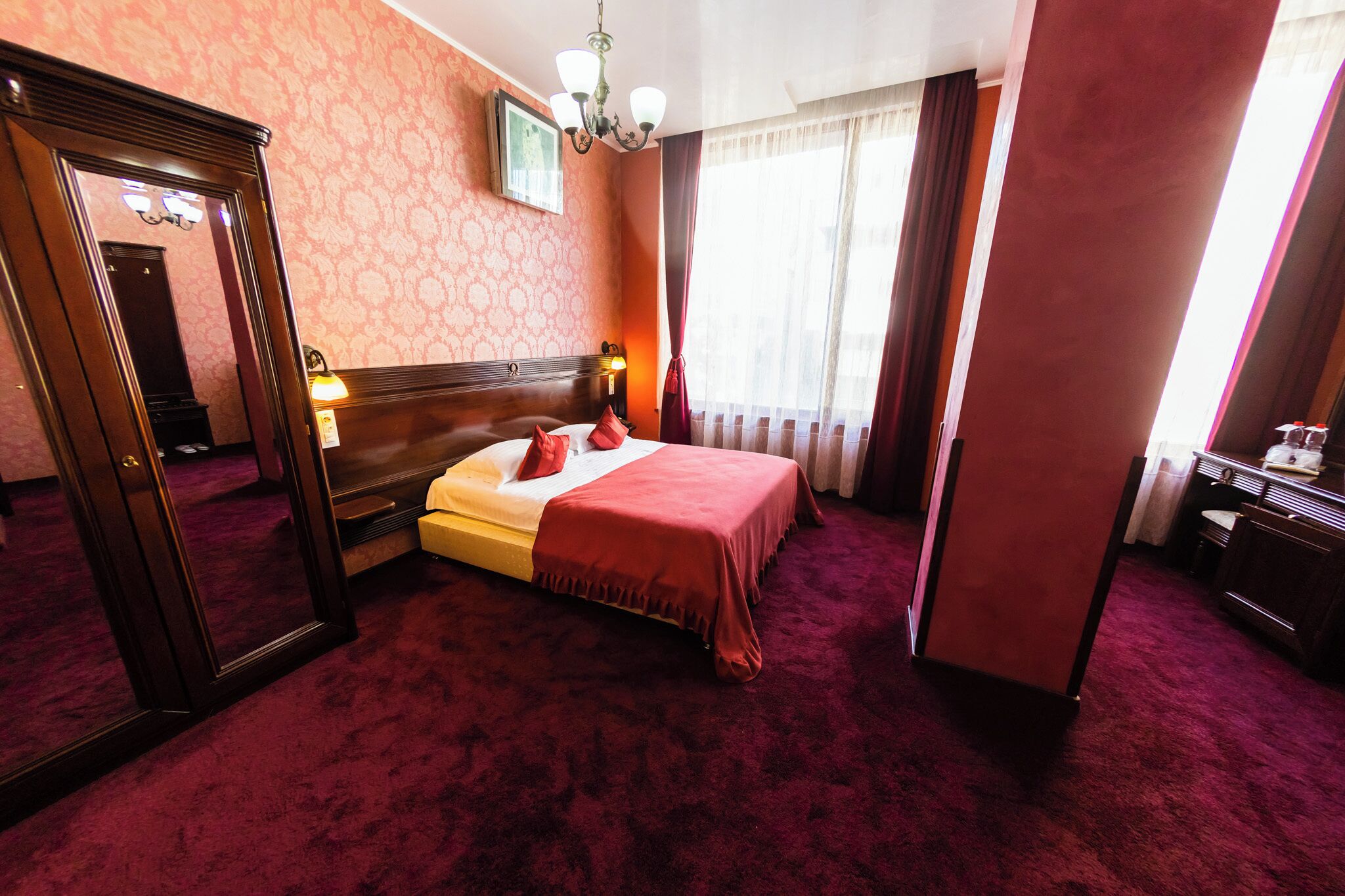 Ein Hotelzimmer mit einem großen Bett, einem Spiegel, einer Deckenleuchte und einem Fenster mit Vorhängen.