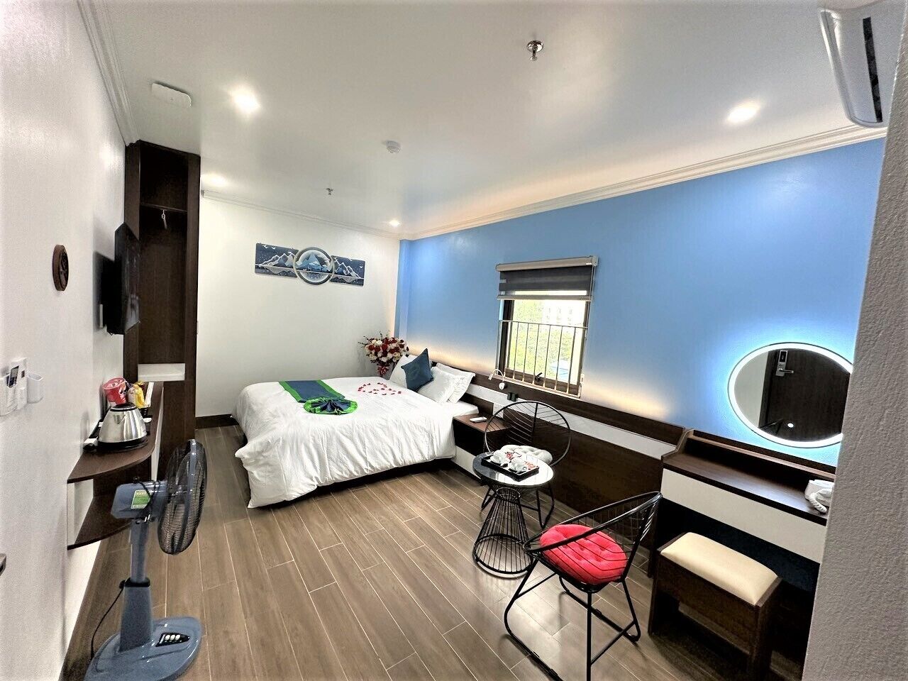 A compact hotel room with a bed, a small desk with a chair, a vanity with a mirror, and a window with blinds.