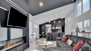 A modern living room with a flat-screen TV, fireplace, grey sofa with red and black pillows, dining area with chairs, and a kitchen in the background.