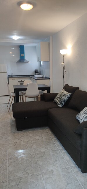 A modern living room with a dark brown sectional sofa, a dining table with chairs, and a kitchen in the background. - APARTAMENTO ENTRE SALINES Y PLAYA (Calpe)