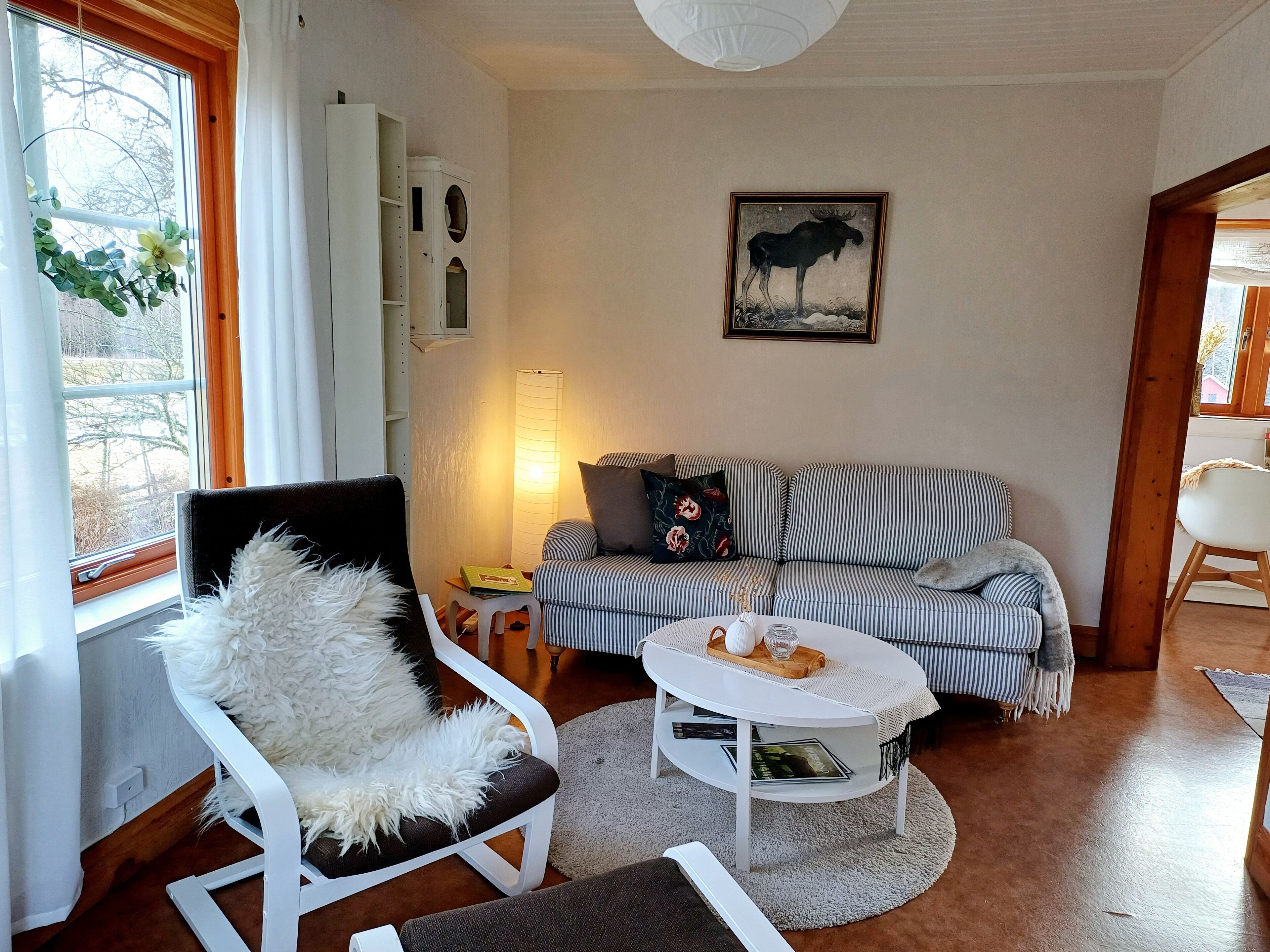 Un salon chaleureux avec un canapé à rayures, un fauteuil blanc doté d’un coussin moelleux, une table basse ronde ornée d’un vase et de livres, une lampe, et un tableau représentant un élan, accroché au mur.