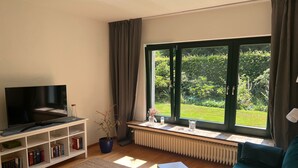 Un soggiorno con televisione, libreria, una pianta in vaso e vista su un giardino attraverso una grande finestra.