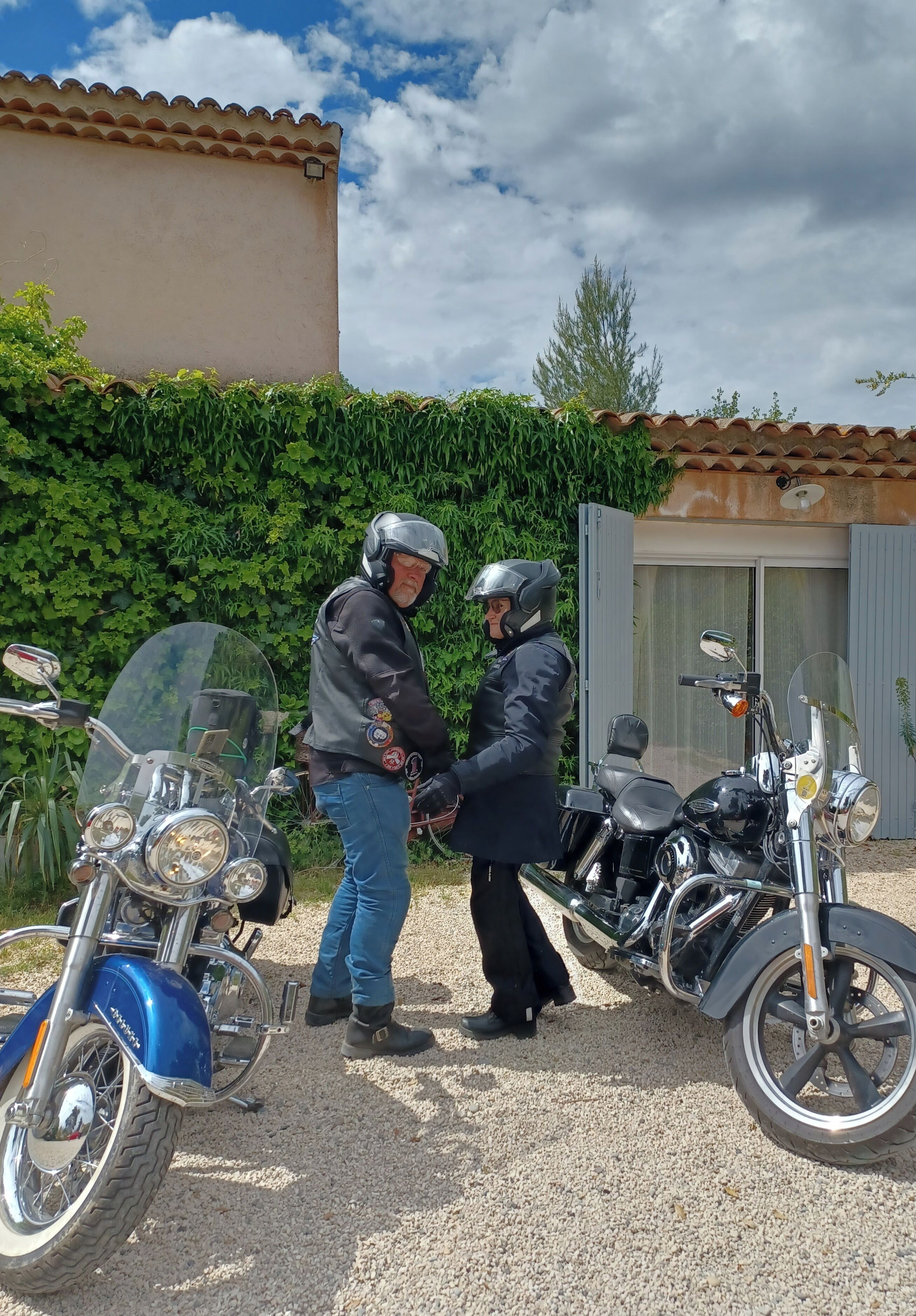 Dos motocicletas estacionadas sobre una superficie de grava, con un edificio y vegetación al fondo.