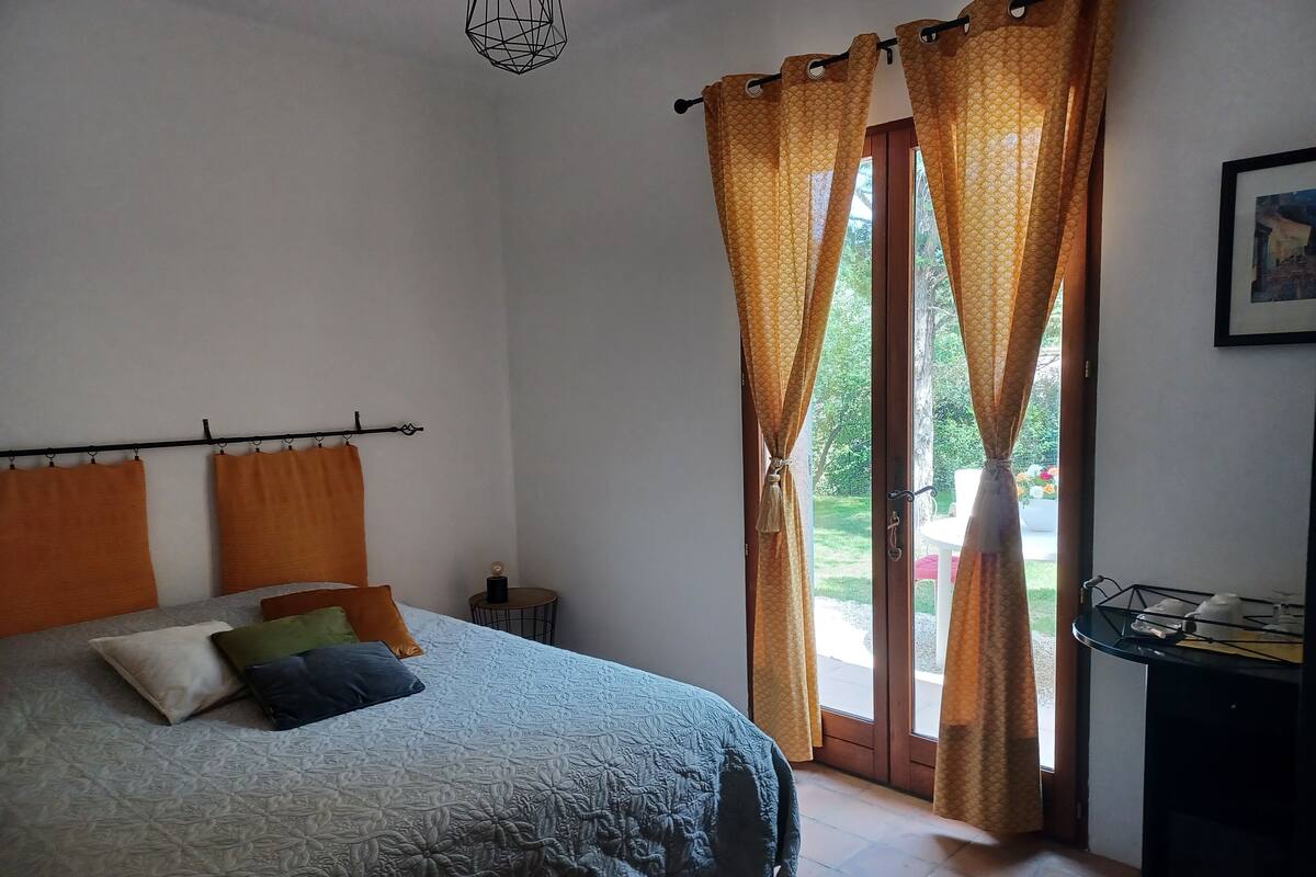A bedroom with a bed, orange curtains, a small table, and a view of the outdoors.