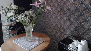 Een tafel met een glazen vaas gevuld met bloemen, behang met een patroon, en een dienblad met witte kopjes en schotels.