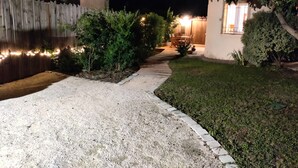 A nighttime view of a residential area with a paved walkway, a wooden fence, and a small garden area with a table and chairs.