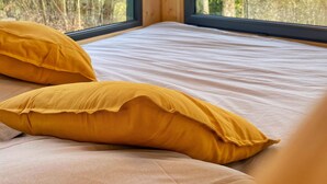 Habitación con paredes de madera y ventanas amplias que dan a un bosque. Se observa una cama con ropa blanca y dos almohadas amarillas.