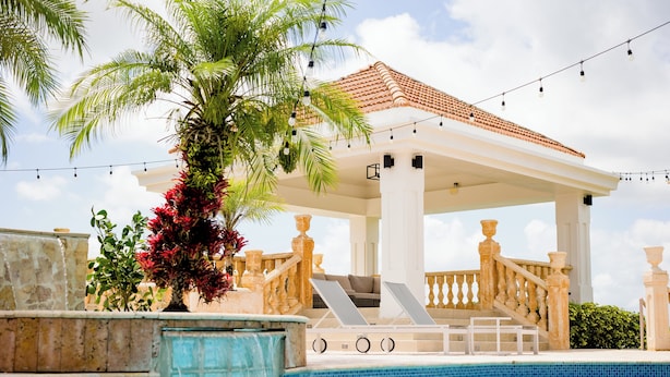 Un área de piscina con zona de estar cubierta, una palmera y una piscina con azulejos.