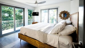 Un dormitorio con una cama grande, un ventilador de techo, un televisor en la pared y un balcón con vistas a la vegetación.