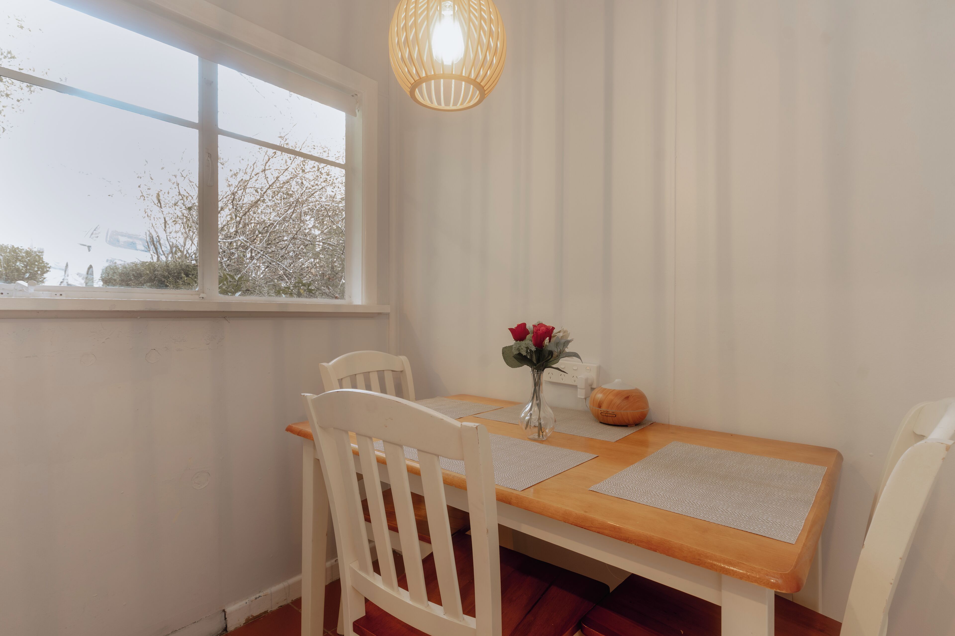Een eetruimte met een houten tafel, witte stoelen, een vaas met rode bloemen en een hanglamp.