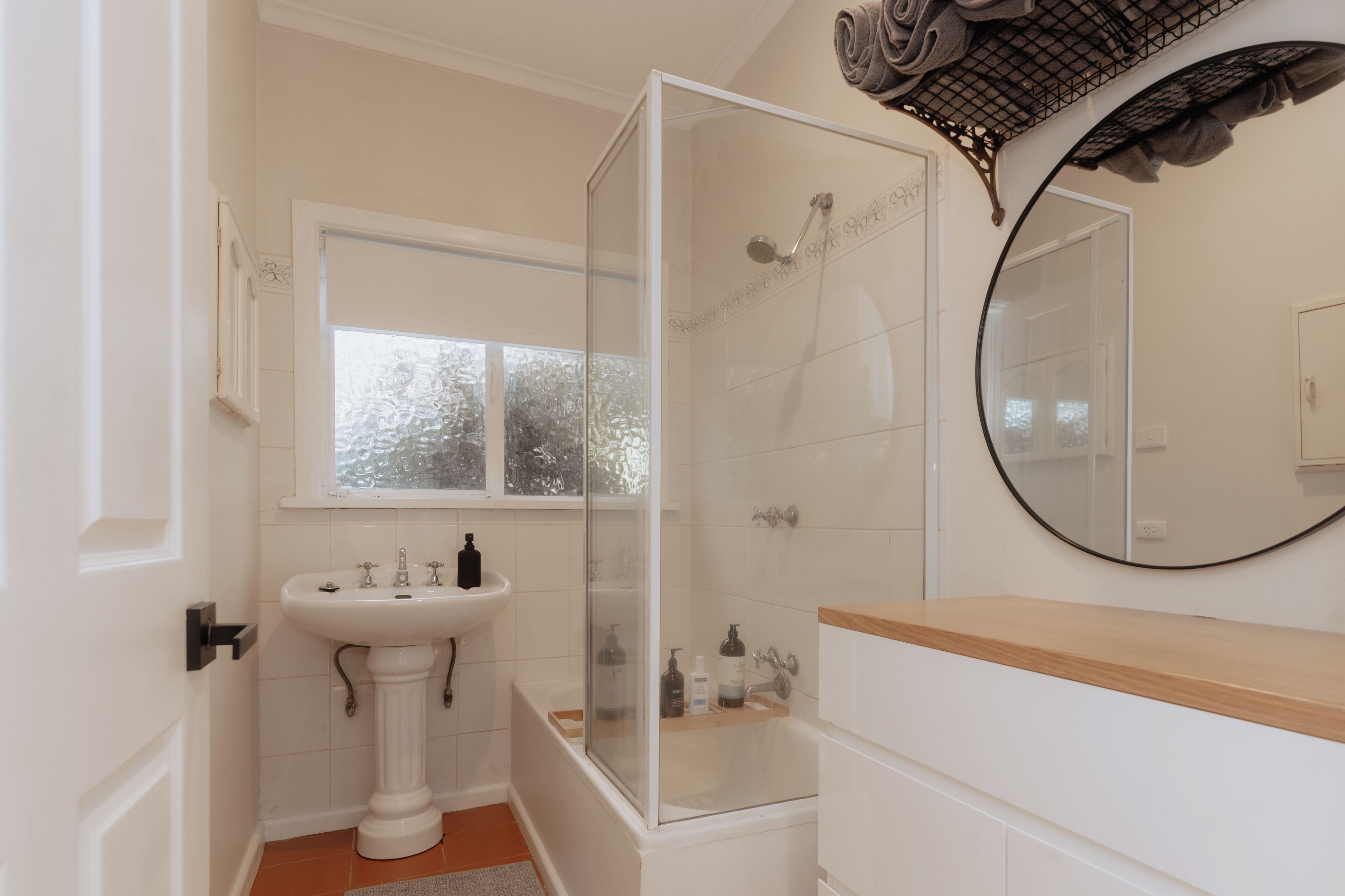 A bathroom with a pedestal sink, a bathtub, a glass-enclosed shower, and a round mirror.