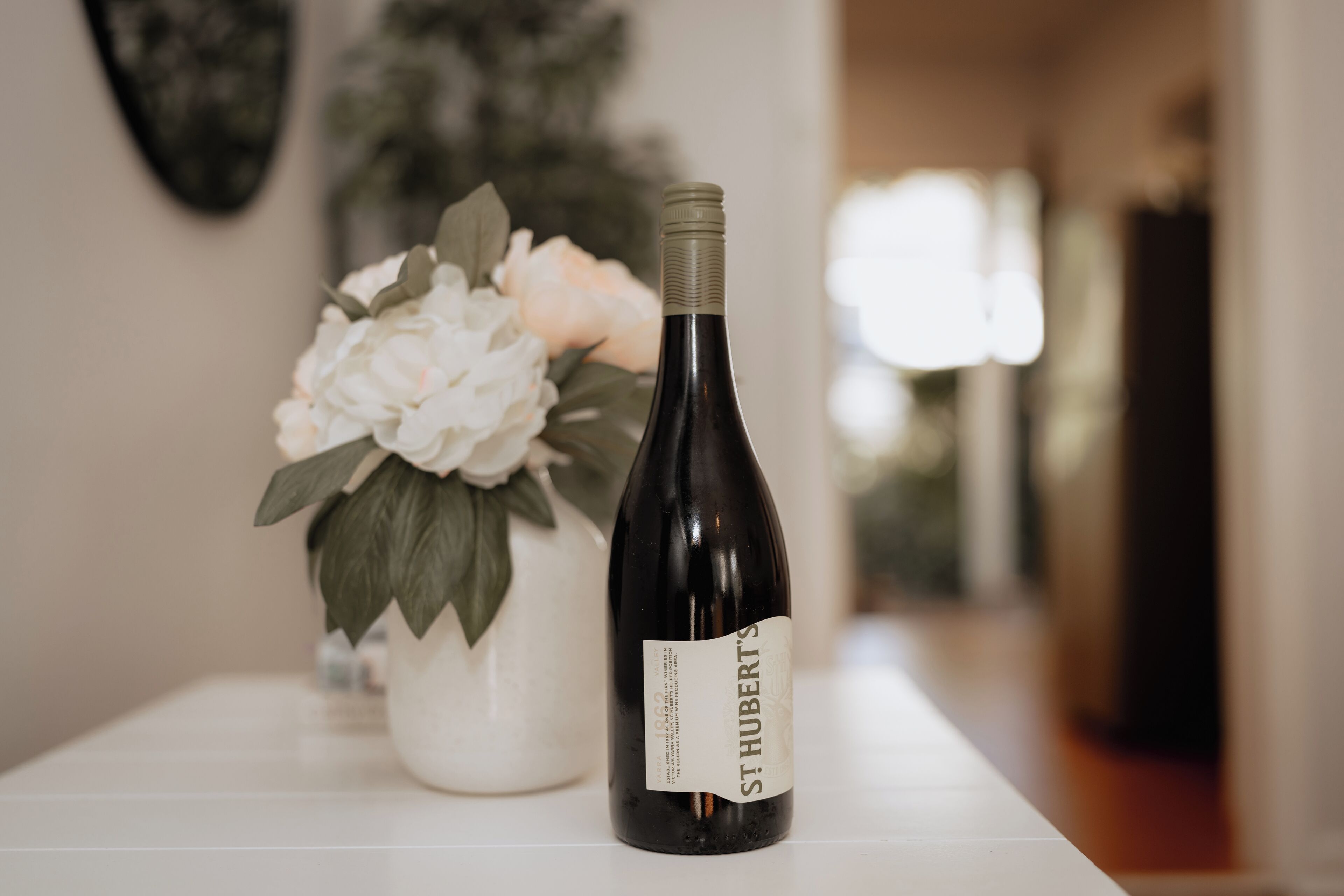 Una botella de vino St. Hubert sobre una mesa blanca, con un jarrón blanco lleno de flores blancas al fondo.
