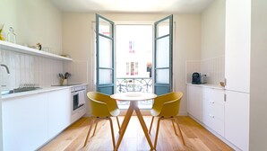 Une cuisine compacte avec des meubles blancs, un plancher en bois, une table à manger avec deux chaises jaunes et, par des portes ouvertes, une vue sur un balcon.