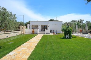 A modern villa with a paved driveway leading to a white entrance, a small patio with outdoor seating, and a well-maintained lawn.