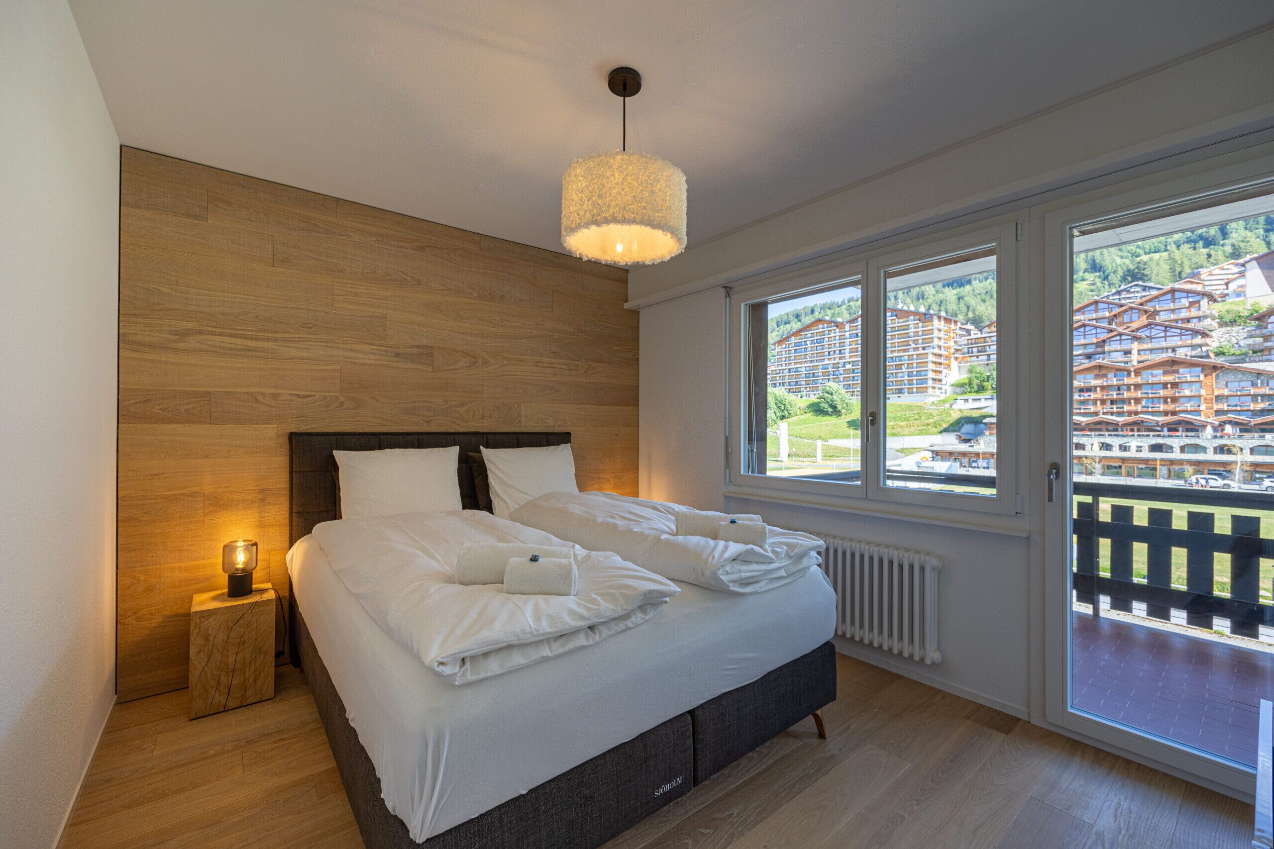 A modern bedroom with a wooden headboard, a large bed with white linens, a bedside table with a lamp, and a view of a building complex through large windows.