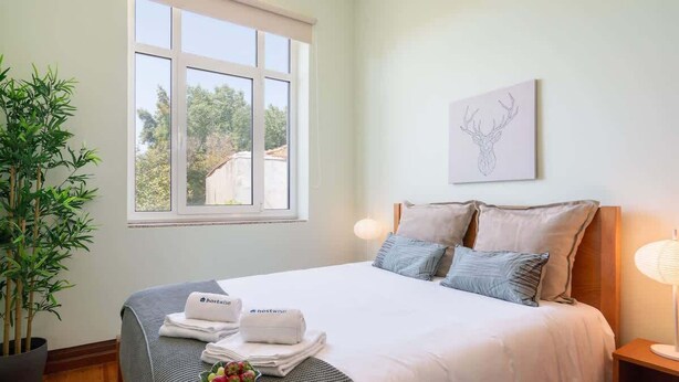 Uma cama bem arrumada com manta branca e cinza, cabeceira com dois travesseiros, mesa de cabeceira com abajur e uma janela com vista para árvores e edifícios.