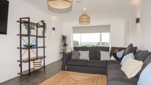 A modern living room with a dark grey sectional sofa, a wooden shelving unit, a large window, and a hanging woven light fixture. - Dolwyn (Menai Bridge)