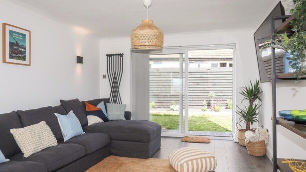 A modern living room with a dark grey sectional sofa, a woven floor lamp, and a glass door leading to a balcony with a view of a garden and a wooden fence. - Dolwyn (Menai Bridge)
