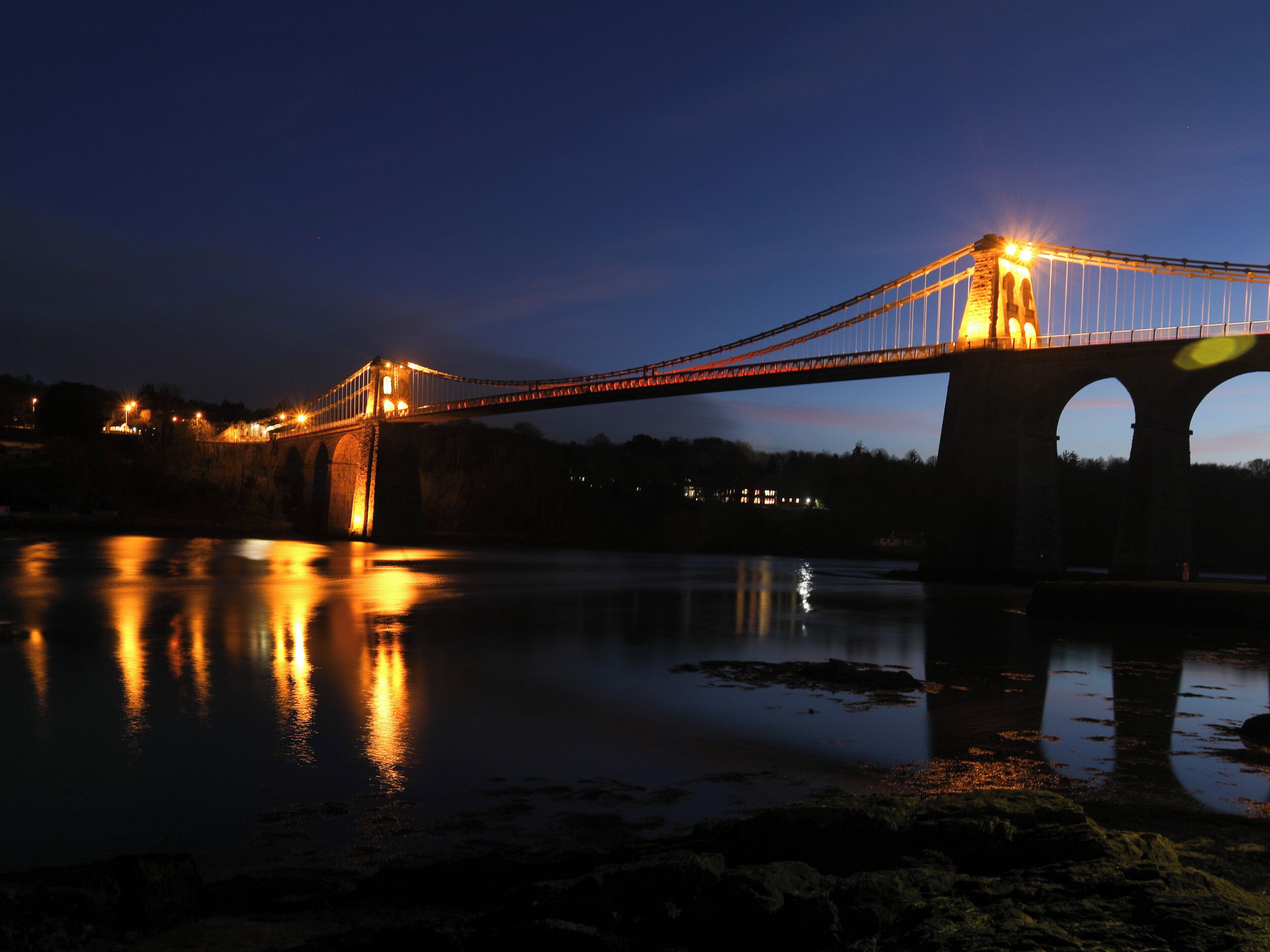Vista nocturna de un puente con luces iluminadas reflejándose en el agua.