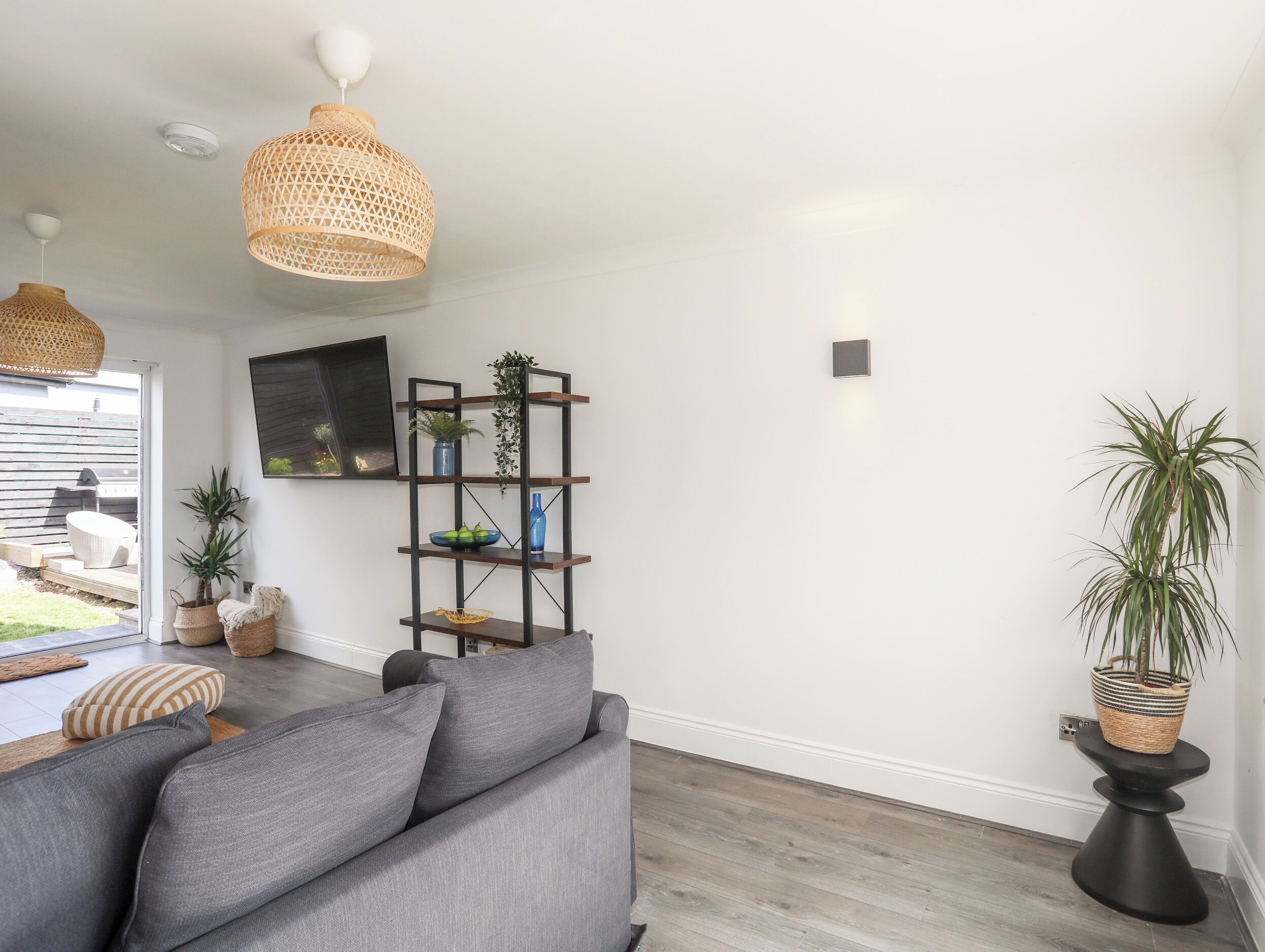 Una sala moderna con un sofá gris, piso de madera, televisor montado en la pared y una estantería con plantas y adornos.