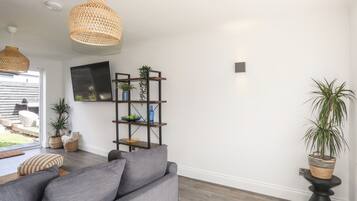 Una sala moderna con un sofá gris, piso de madera, televisor montado en la pared y una estantería con plantas y adornos.