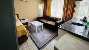 A compact hotel room with a sofa, a small white table, a kitchen area with a sink, and a window with curtains. - PALVAN ACCOMMODATION (Antalya)