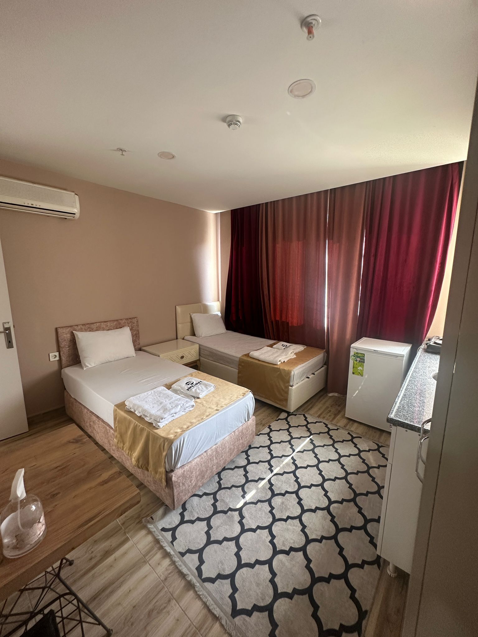 Een hotelkamer met twee bedden, een klein keukentje, een kamerkleed met patroon en een airconditioning.