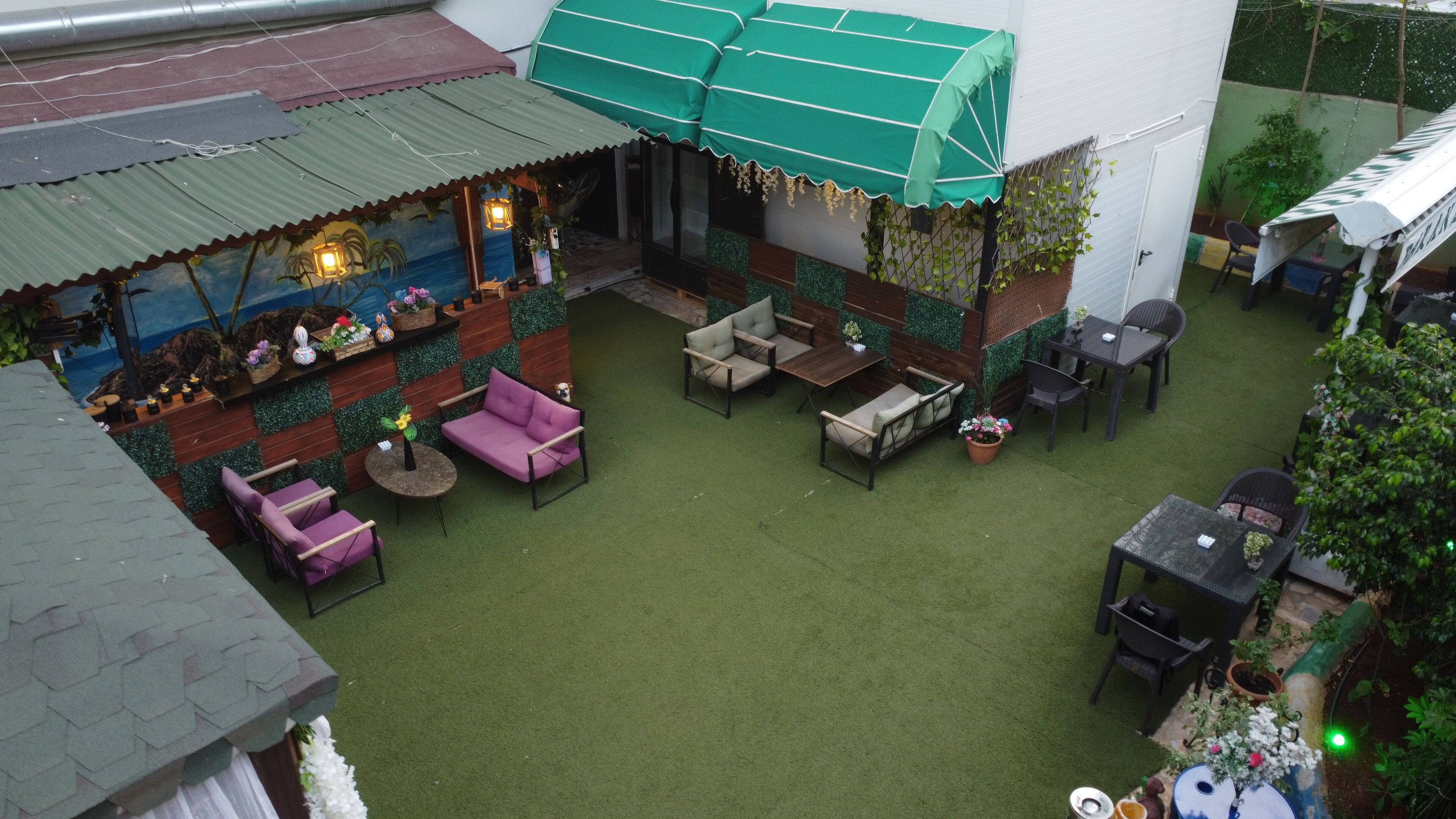 An outdoor patio area with green artificial grass, a covered seating area with cushions, and a variety of potted plants.
