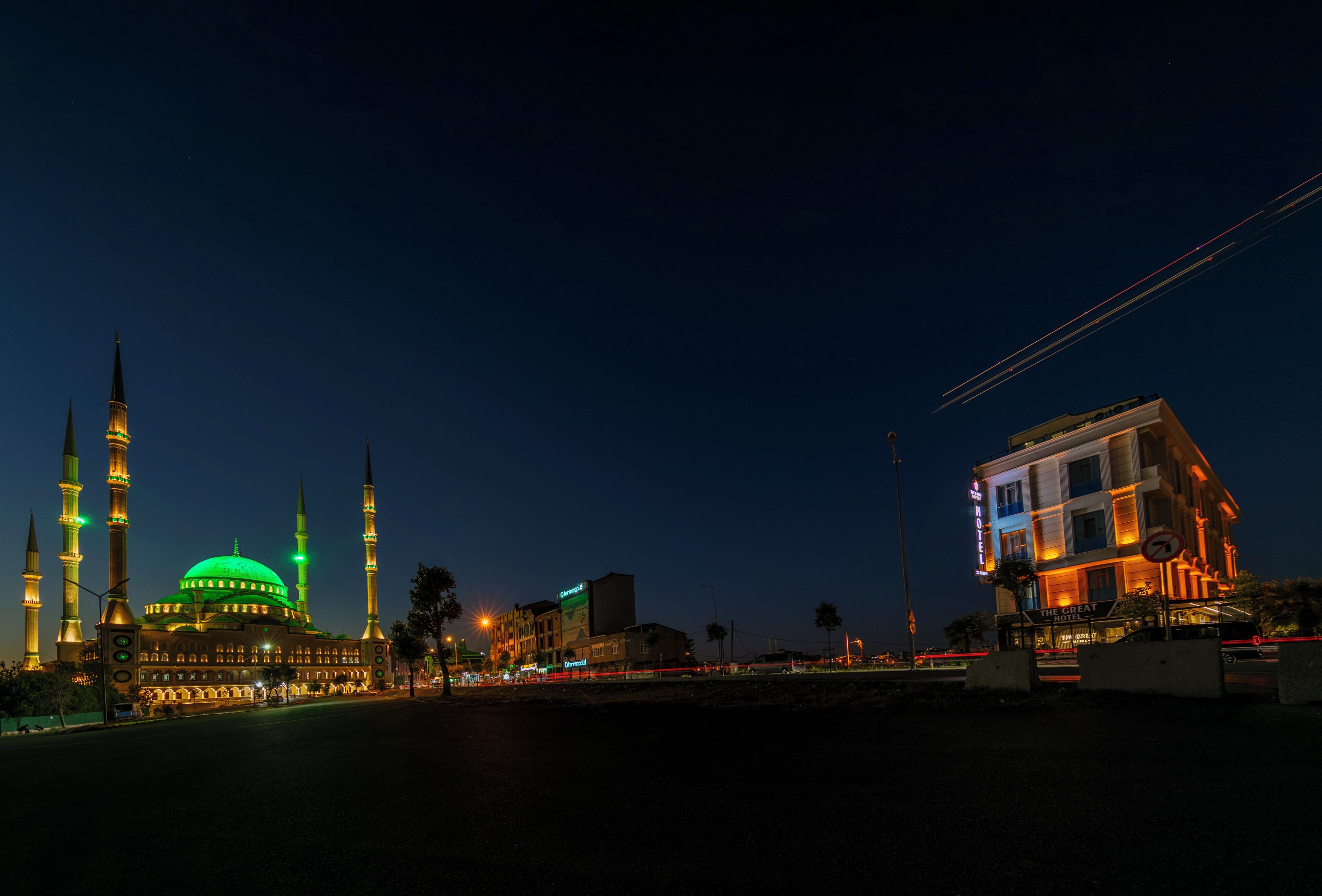 Een moskee met meerdere minaretten en een groen koepeltje, naast een modern gebouw met een bord waarop staat: "The Great Hotel".