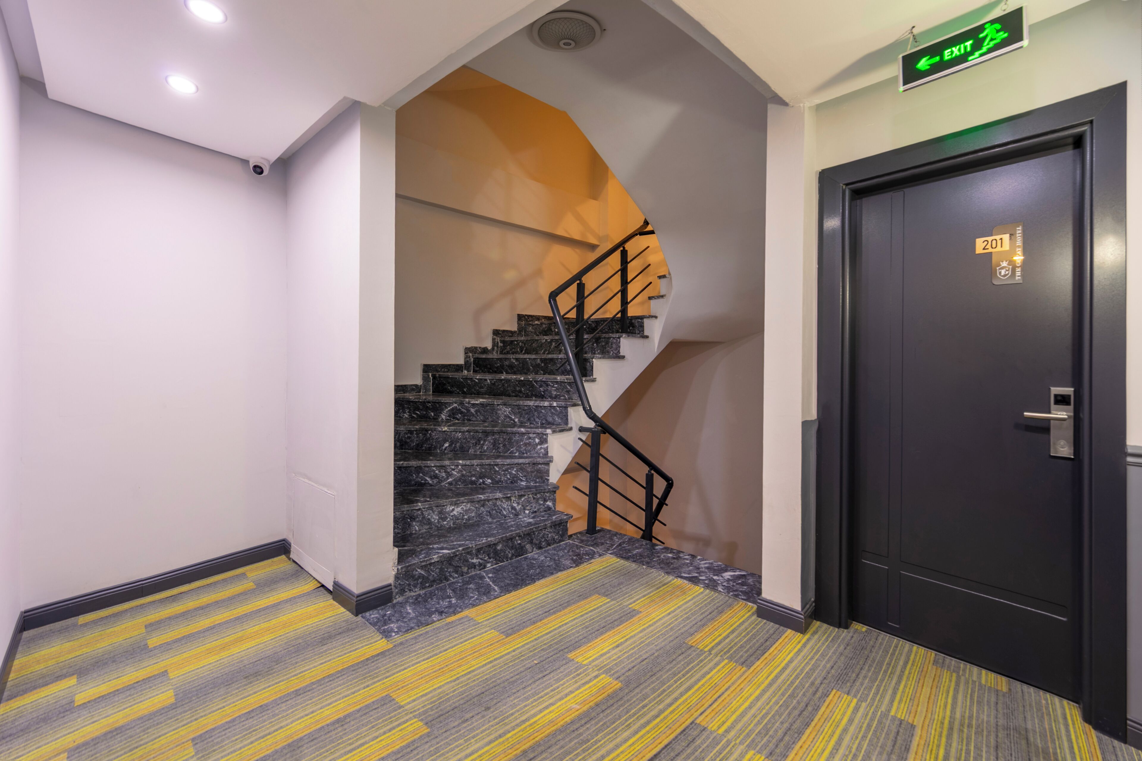 A hotel corridor with a marble staircase, a black door with a number plate, and a yellow and grey patterned carpet.