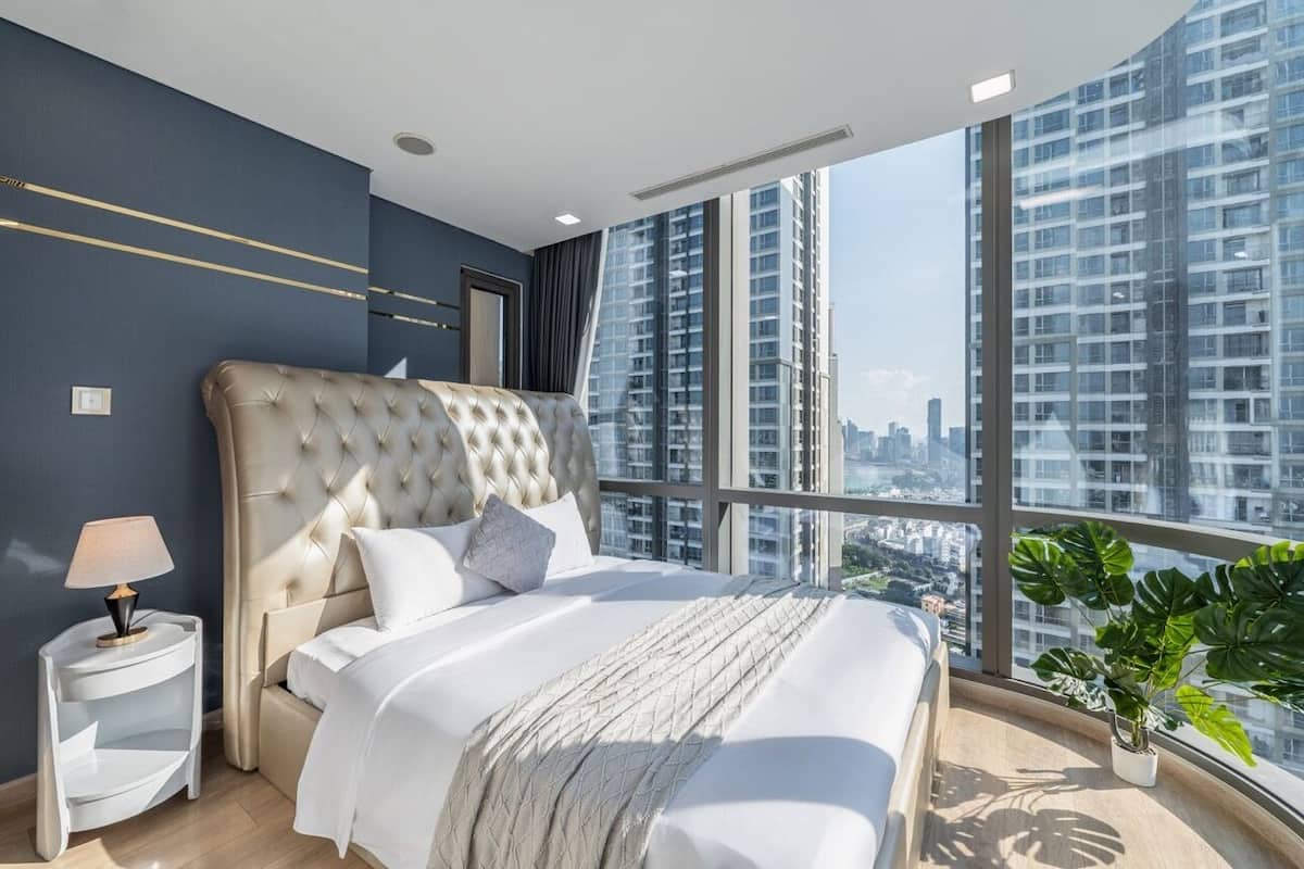 A modern bedroom with a large bed, a nightstand, a potted plant, and a city view through large windows.