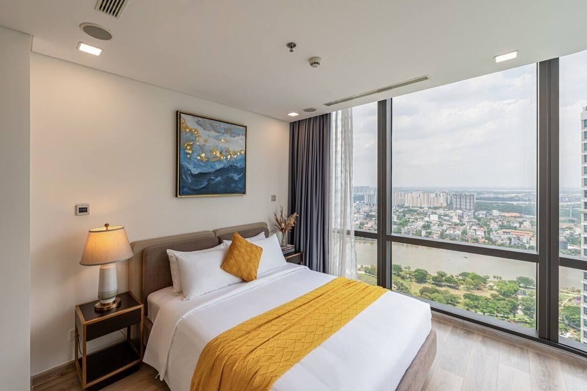 A hotel room with a large window, a bed, a nightstand, a lamp, and a painting on the wall.