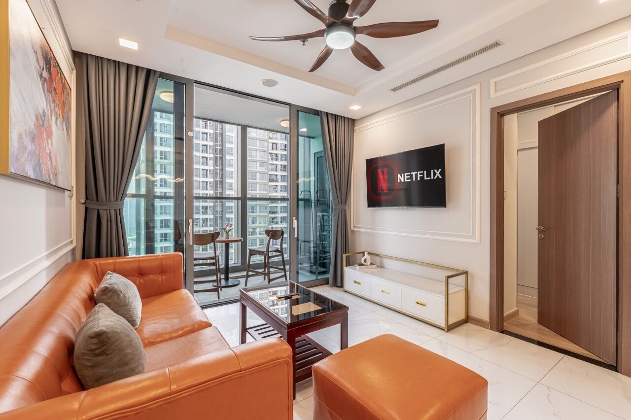 A modern living room with a leather sofa, a glass coffee table, a ceiling fan, a TV mounted on the wall, and a large window with curtains.