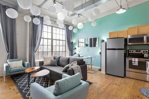 A modern living room with grey sofas, a coffee table, and a kitchen area with stainless steel appliances. - Loft Living in the Trendy Warehouse District (Durham)