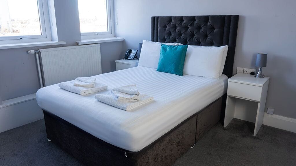 Una cama bien hecha con sábanas blancas y una almohada azul turquesa, un teléfono en la mesita de noche y una lámpara.