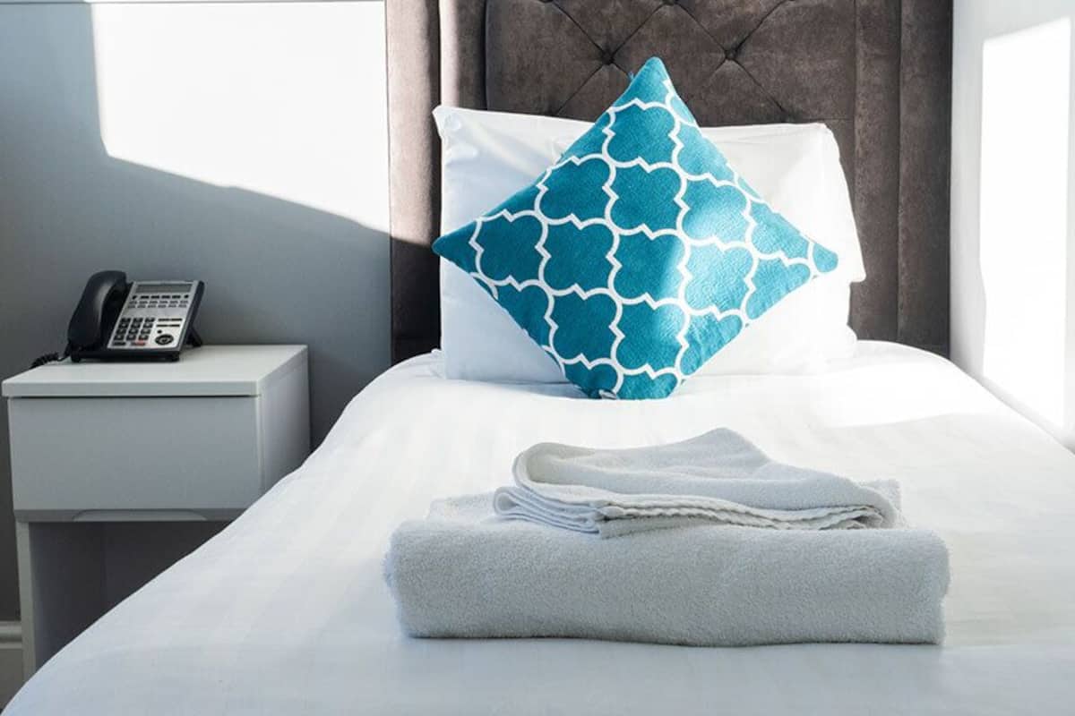 A neatly made bed with a folded towel, a patterned pillow, a bedside phone, and a nightstand.