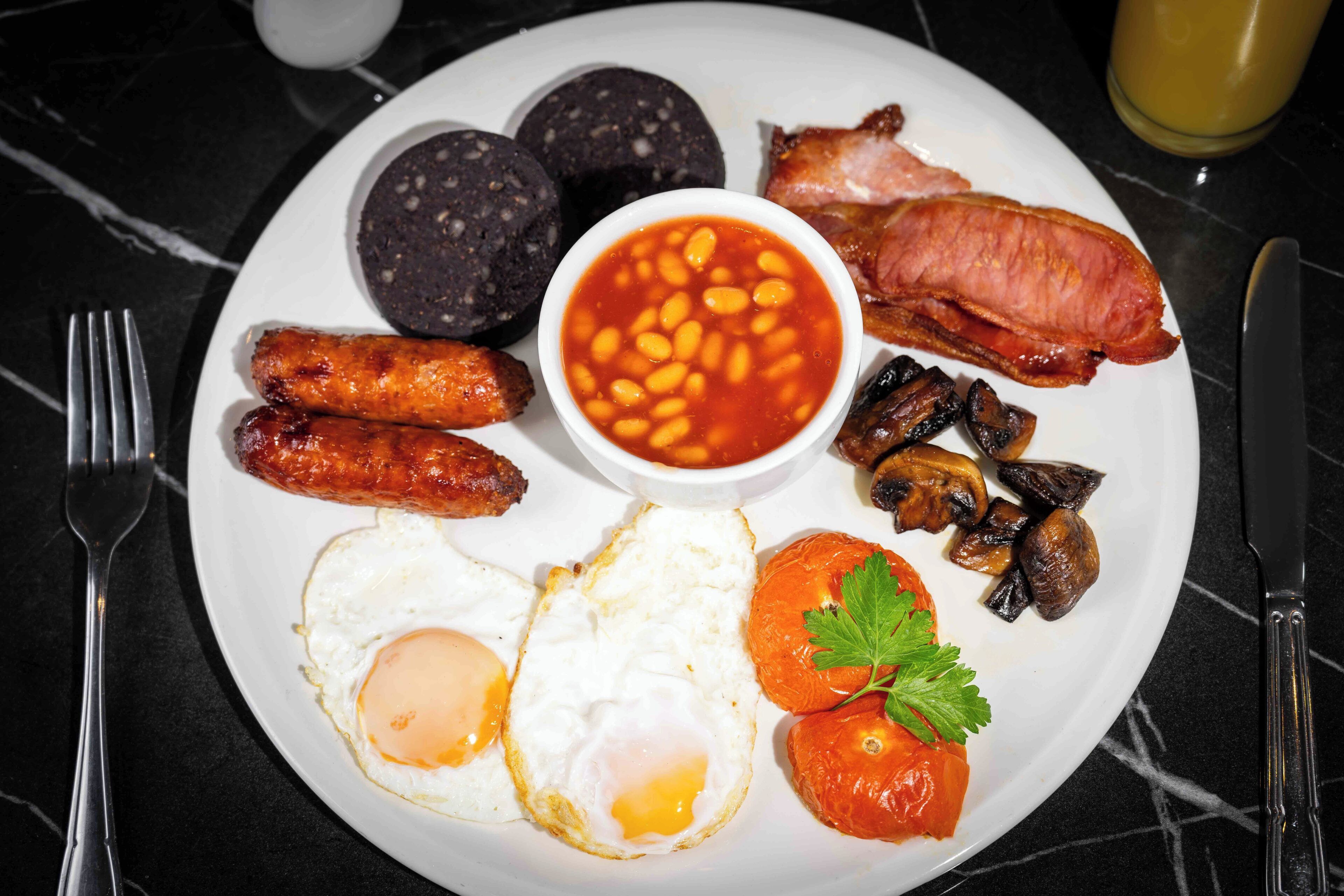 A plate of breakfast food including sausages, eggs, beans, bacon, mushrooms, and a tomato.
