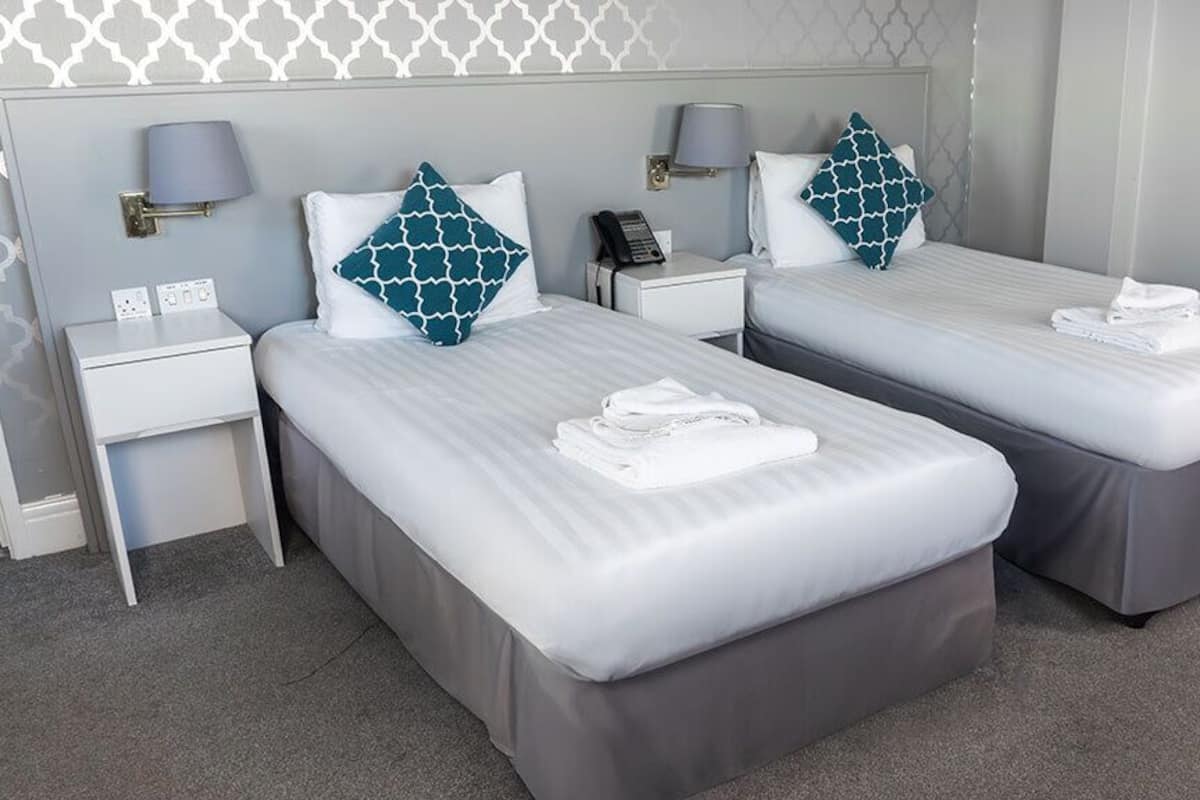 A hotel room with two single beds, each with a pillow and a folded towel. There is a bedside table with a lamp and a phone. The room has a grey carpet and a patterned wall.