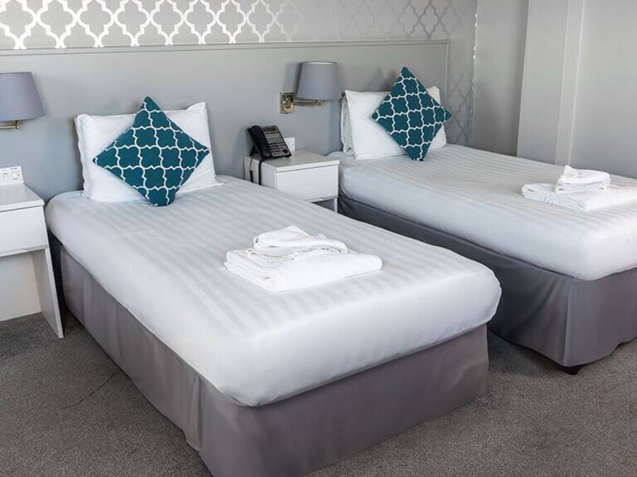 A hotel room with two single beds, each with a pillow and a folded towel. There is a bedside table with a lamp and a phone. The room has a grey carpet and a patterned wall.