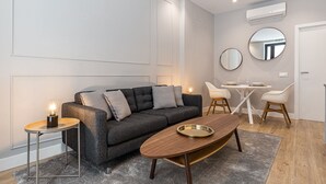 Ein modernes Wohnzimmer mit einem dunkelgrauen Sofa, einem runden Couchtisch und einem Esstisch mit Stühlen. Zwei Spiegel sind an der Wand montiert, und es gibt eine Klimaanlage.