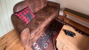 Un salon chaleureux avec un canapé en cuir marron, un tapis à motifs, un banc en bois, un morceau de fourrure décorative accroché au mur, une bibliothèque et un miroir rond.