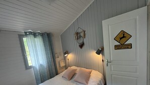 Une chambre à coucher avec un lit, deux lampes de chevet, une fenêtre avec des rideaux, une porte ornée d’un panneau d’avertissement indiquant un passage de cerfs, et un attrape-rêves fixé au mur.
