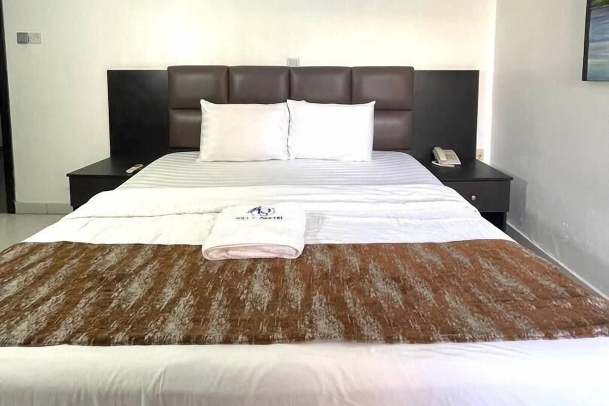 A modern hotel room with a large bed, a dark headboard, white bedding, and a brown patterned throw.
