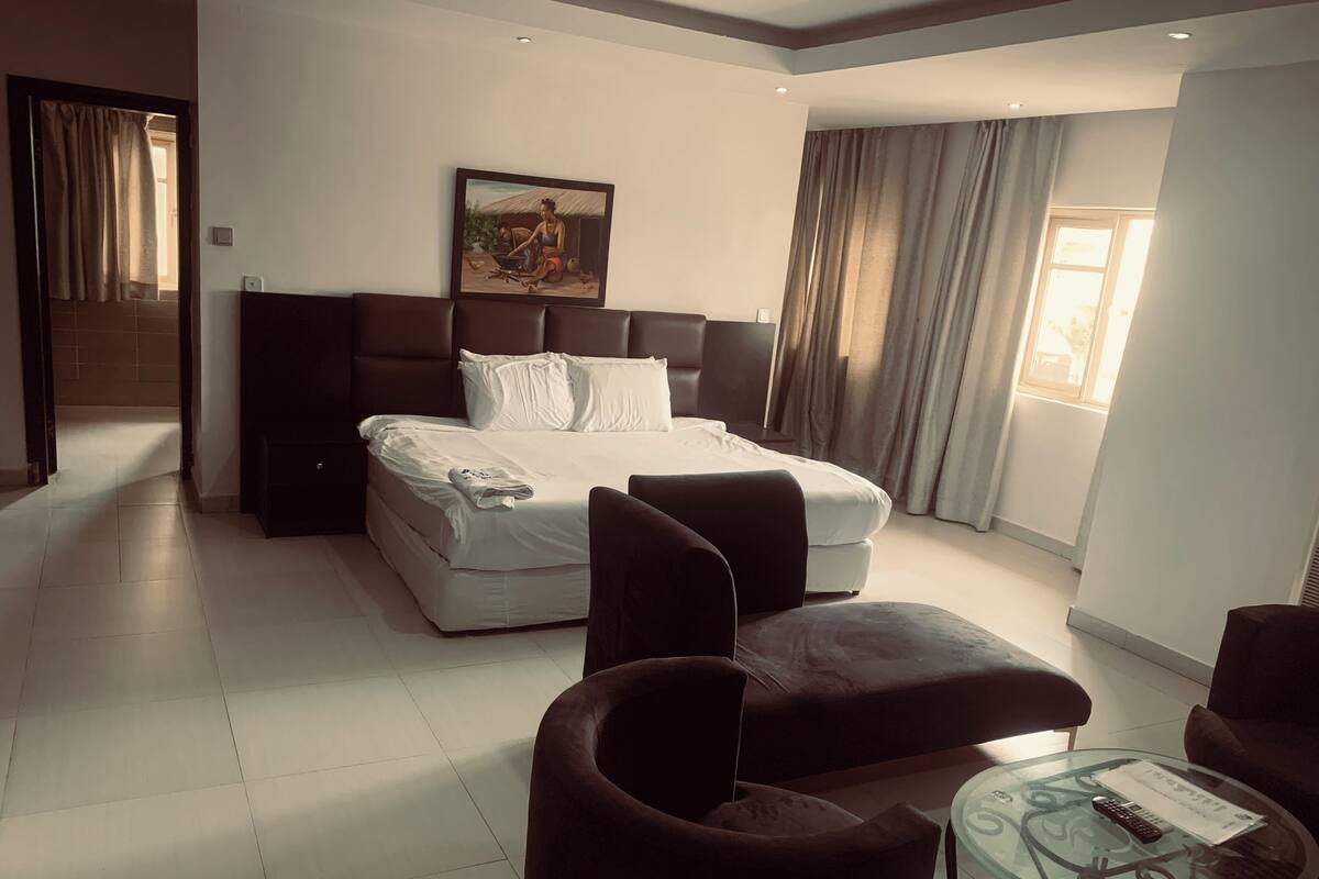 A modern hotel room with a large bed, a seating area with two armchairs, a glass coffee table, and a painting on the wall.
