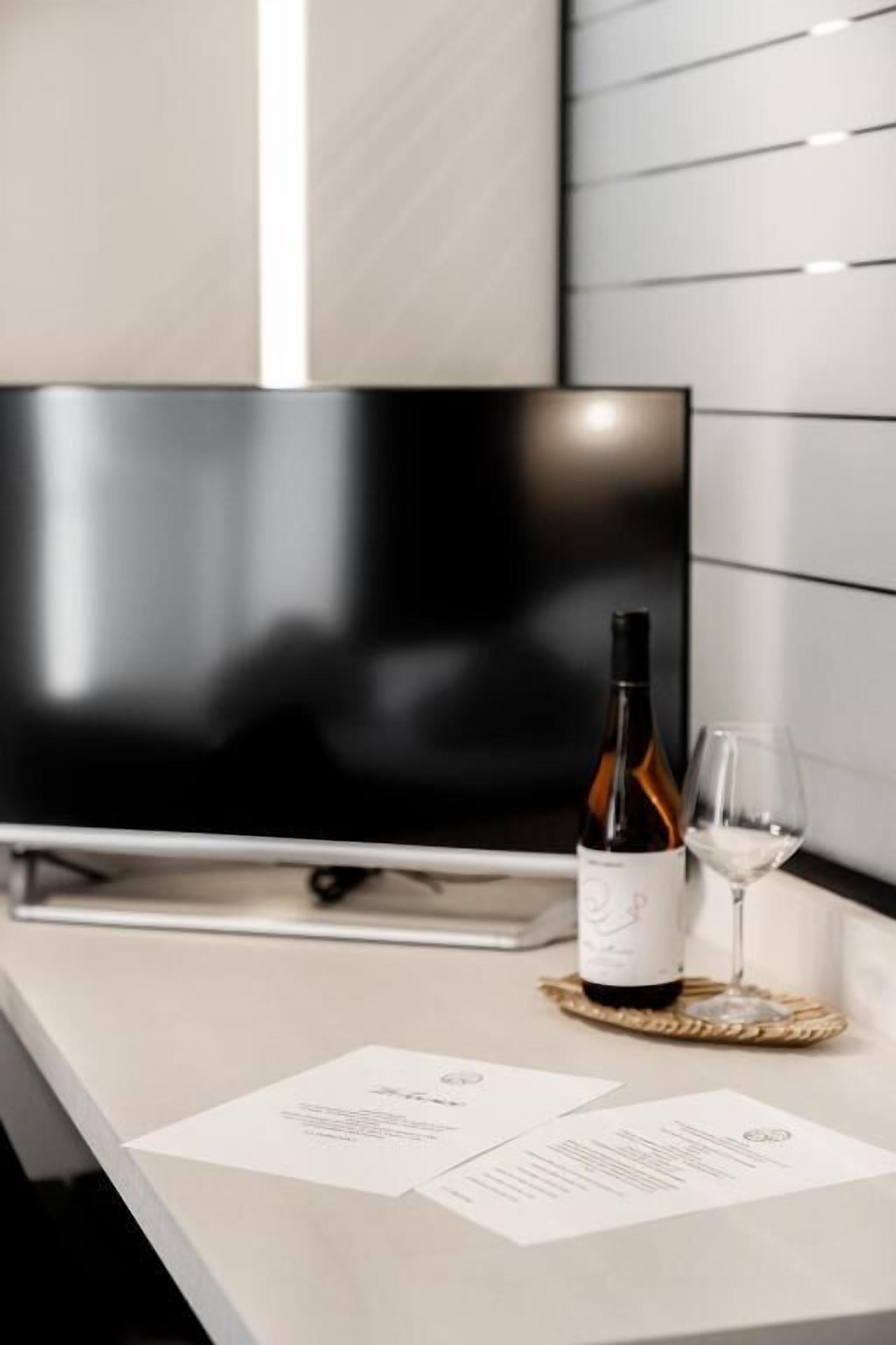 A modern hotel room with a flat-screen TV, a bottle of wine, and a glass on a wooden coaster.