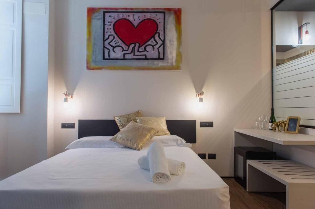 A hotel room with a bed, pillows, a towel, a wall art piece, a bedside table, and a mirror.