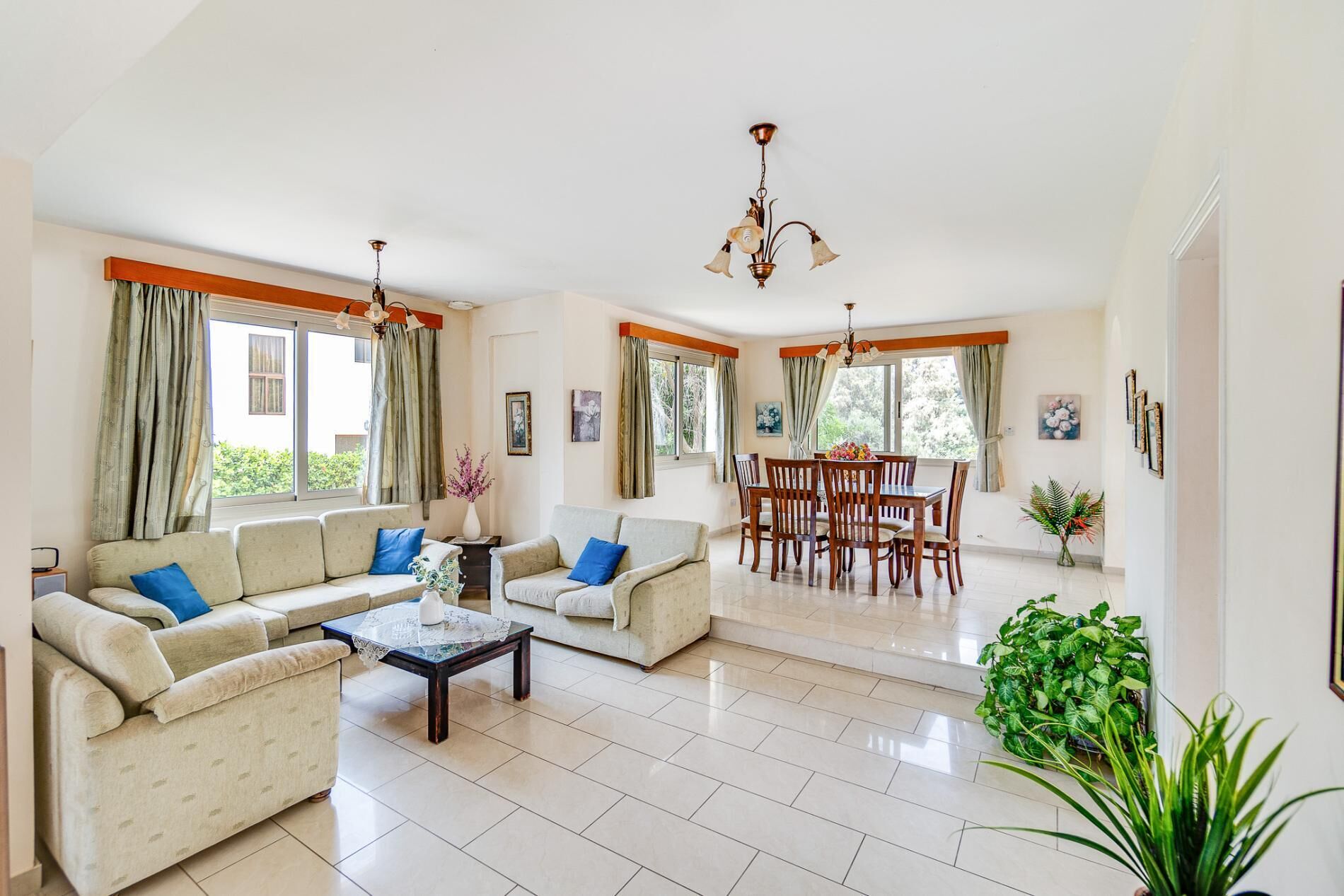 A spacious living room with a sofa set, a dining table, and chairs. There are large windows with curtains, a chandelier, and a potted plant.