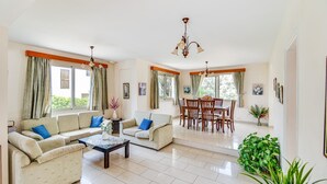 Amplia sala de estar con un conjunto de sofás, una mesa de comedor y sillas. Cuenta con ventanas grandes con cortinas, una lámpara de araña y una planta en maceta.