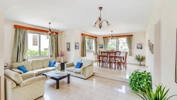 A spacious living room with a sofa set, a dining table, and chairs. There are large windows with curtains, a chandelier, and a potted plant.